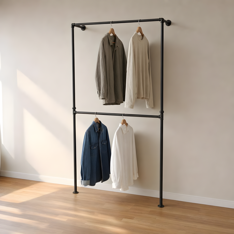 Portant de penderie sol-mur à deux barres en tuyaux de plomberie noirs Ø 27 mm, vêtements sur cintres en bois, mur beige et parquet chêne
